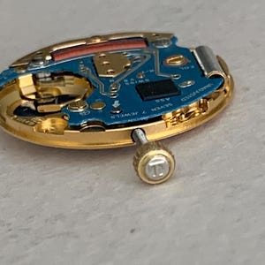 May include: Close-up of a disassembled watch movement. The watch components include a gold-toned case, a blue circuit board with small electronic components, and a screw. The watch movement is labeled "SWISS 7 JEWELS".