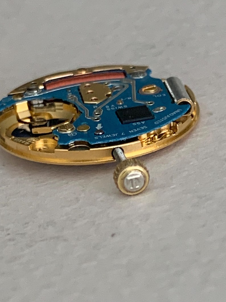 May include: Close-up of a disassembled watch movement. The watch components include a gold-toned case, a blue circuit board with small electronic components, and a screw. The watch movement is labeled "SWISS 7 JEWELS".