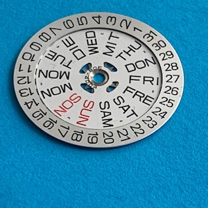 May include: A silver watch calendar dial with days of the week and numbered dates. The days of the week are in English, with "SUN" in red. The dial is on a blue background.