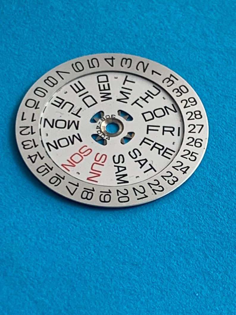 May include: A silver watch calendar dial with days of the week and numbered dates. The days of the week are in English, with "SUN" in red. The dial is on a blue background.