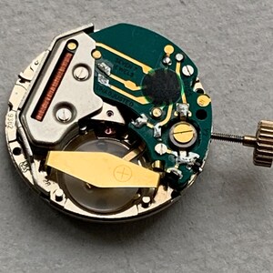 Puede incluir: Primer plano de un movimiento de reloj con un rotor dorado y una placa de circuito verde. La placa tiene el texto "SEVEN JEWELS" y "UNADJUSTED". El movimiento es circular con una corona dorada.