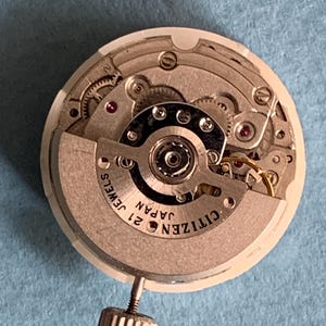 May include: A close-up of a Citizen watch movement with intricate gears and components. The watch mechanism is silver and gold-toned, with the text "CITIZEN JAPAN 21 JEWELS" visible. A small, textured silver crown is attached.