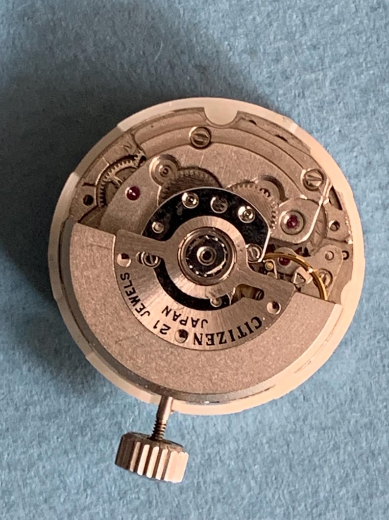May include: A close-up of a Citizen watch movement with intricate gears and components. The watch mechanism is silver and gold-toned, with the text "CITIZEN JAPAN 21 JEWELS" visible. A small, textured silver crown is attached.
