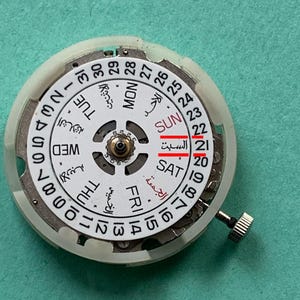 May include: A close-up of a watch movement with a white dial and a silver-colored metal casing. The dial displays the days of the week in English and Arabic, with the date highlighted. The watch crown is visible on the right side.