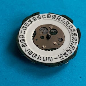 May include: A watch movement with a white dial displaying black numerals from 1 to 31, representing the days of the month. It has a green outer ring and a central mechanism with small gears and screws. The image is set against a blue background.