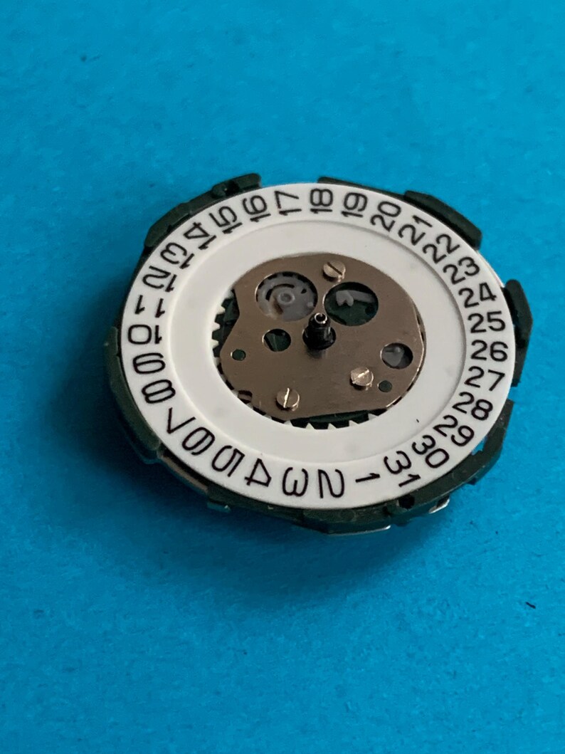May include: A watch movement with a white dial displaying black numerals from 1 to 31, representing the days of the month. It has a green outer ring and a central mechanism with small gears and screws. The image is set against a blue background.