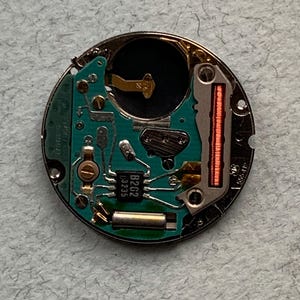 May include: A circular watch movement with a green circuit board and various electronic components. The movement has a silver metal casing and includes a small black chip labeled "B202 13235". A red coil is visible on the right side.