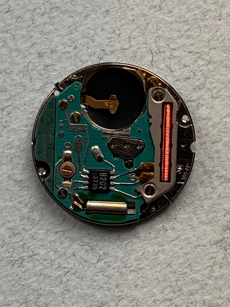 May include: A circular watch movement with a green circuit board and various electronic components. The movement has a silver metal casing and includes a small black chip labeled "B202 13235". A red coil is visible on the right side.