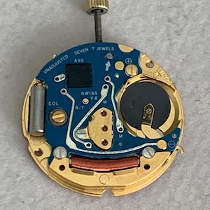May include: A watch movement with a blue circuit board and gold-colored metal components. The text "SEVEN 7 JEWELS" and "SWISS V8" are visible. A battery and a screw are also present.