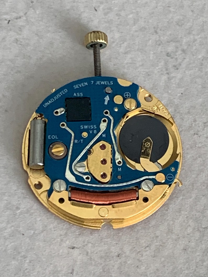May include: A watch movement with a blue circuit board and gold-colored metal components. The text "SEVEN 7 JEWELS" and "SWISS V8" are visible. A battery and a screw are also present.