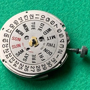 May include: A close-up of a watch movement component with a silver-colored dial. The dial displays the days of the week in English and numbers 1-31. A small, gold-colored component is in the center. A small silver crown is visible.