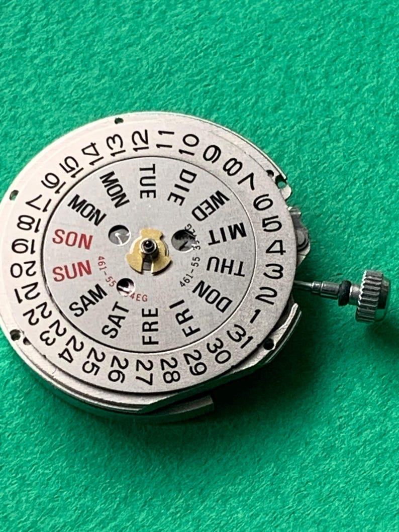 May include: A close-up of a watch movement component with a silver-colored dial. The dial displays the days of the week in English and numbers 1-31. A small, gold-colored component is in the center. A small silver crown is visible.