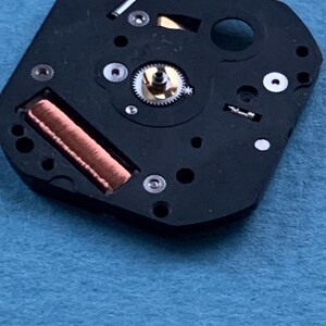 May include: Close-up of a black watch movement with copper coil, silver gears, and small screws. The watch mechanism is set against a blue background. The intricate details of the watch components are visible.