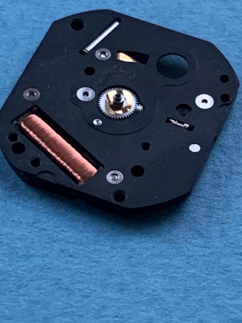 May include: Close-up of a black watch movement with copper coil, silver gears, and small screws. The watch mechanism is set against a blue background. The intricate details of the watch components are visible.