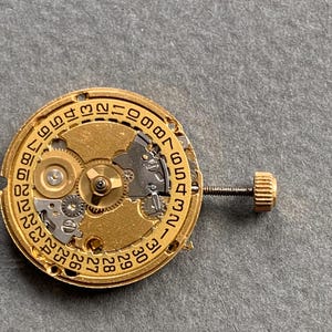 May include: Close-up of a gold-toned watch movement with intricate gears and a date dial. The movement features a screw and a small crown. The background is a neutral gray.