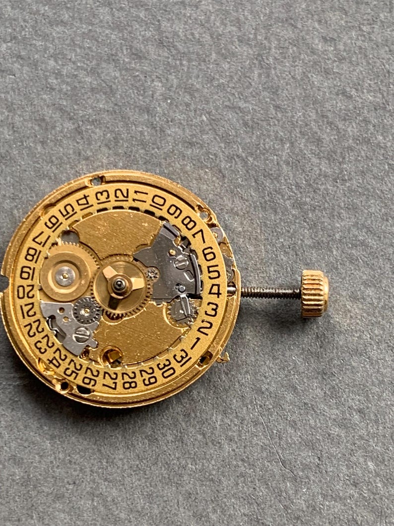 May include: Close-up of a gold-toned watch movement with intricate gears and a date dial. The movement features a screw and a small crown. The background is a neutral gray.