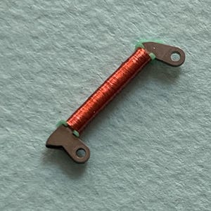 May include: A small, copper-colored coil with dark gray metal end pieces and green insulation. The end pieces have a hole for attachment. The coil is centered on a light blue surface.