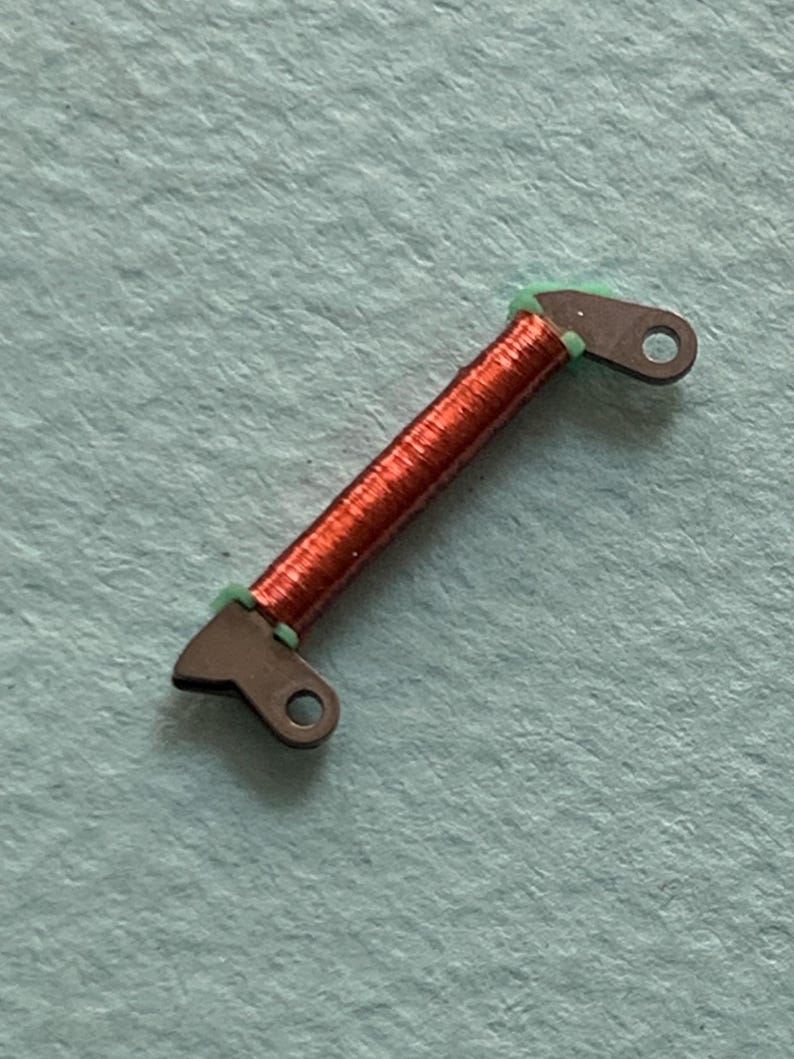 May include: A small, copper-colored coil with dark gray metal end pieces and green insulation. The end pieces have a hole for attachment. The coil is centered on a light blue surface.