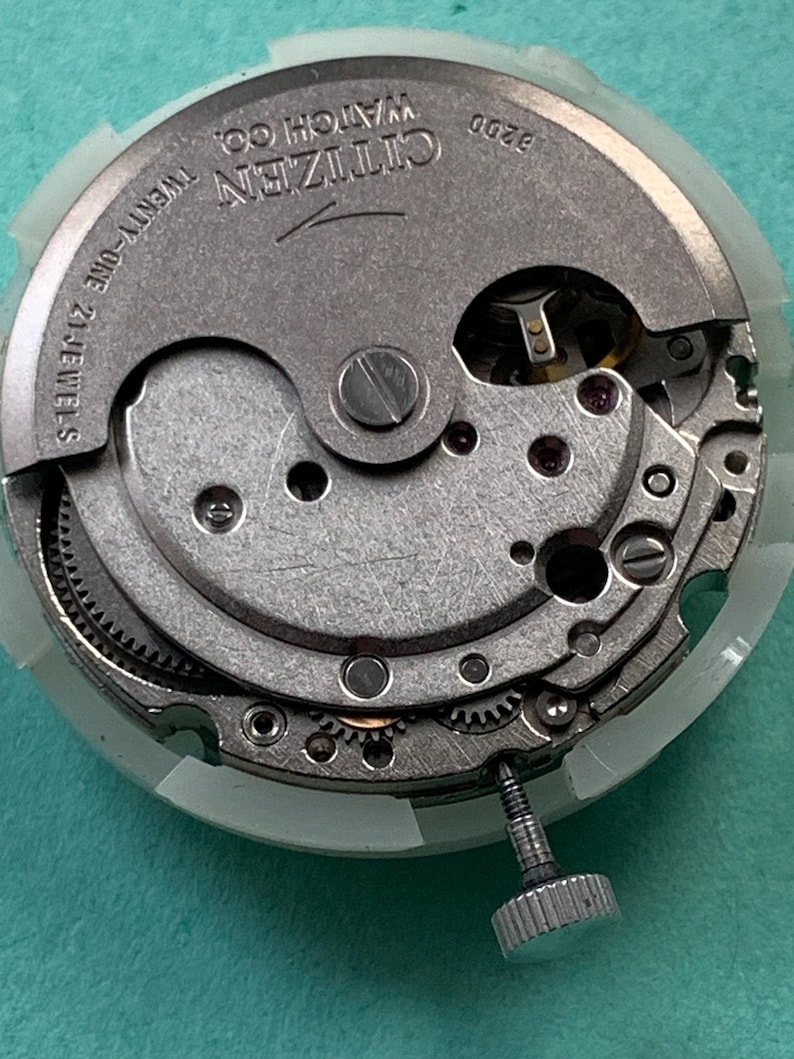 May include: Close-up of a Citizen watch movement, featuring intricate gears, screws, and the text "Citizen Watch Co." and "Twenty-One 21 Jewels." The watch mechanism is silver-toned, with a white plastic ring around the edge. The model number 8200 is also visible.