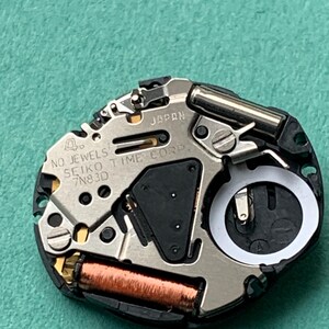May include: Close-up of a watch movement with the text "JAPAN" and "SEIKO TIME CORP". The watch mechanism has a silver metal finish with black and copper-colored components. The watch movement is on a green surface.