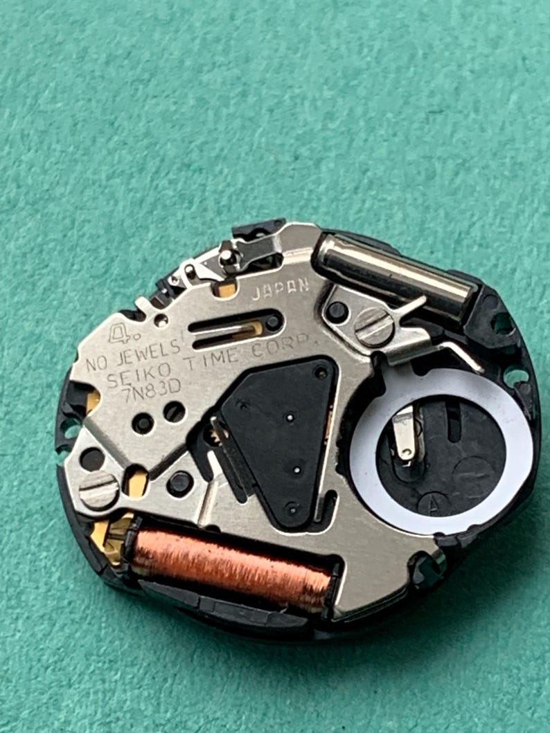 May include: Close-up of a watch movement with the text "JAPAN" and "SEIKO TIME CORP". The watch mechanism has a silver metal finish with black and copper-colored components. The watch movement is on a green surface.