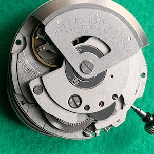 May include: Close-up of a silver-toned Orient watch movement. The mechanism features intricate gears, a balance wheel, and the text "Orient Watch Co. Ltd. Japan". The watch movement is set against a green background.