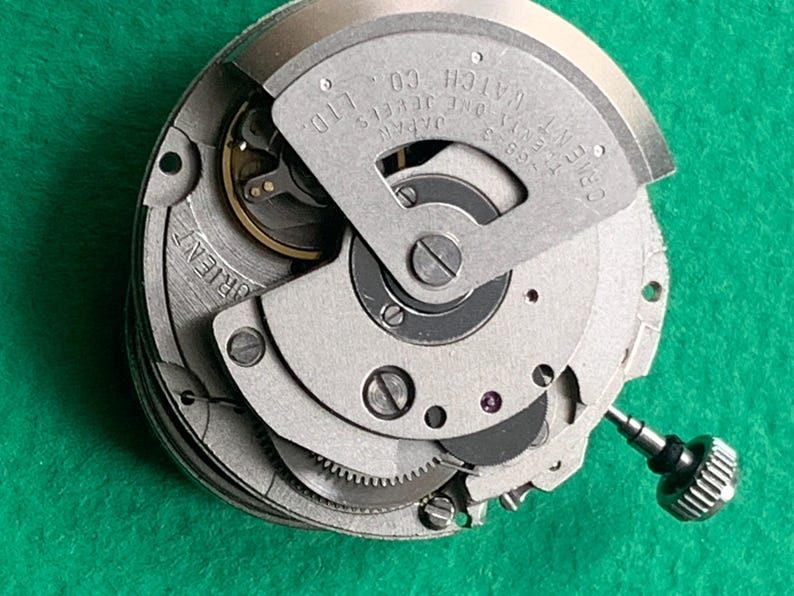 May include: Close-up of a silver-toned Orient watch movement. The mechanism features intricate gears, a balance wheel, and the text "Orient Watch Co. Ltd. Japan". The watch movement is set against a green background.