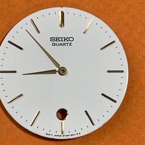 May include: A white Seiko Quartz watch dial featuring gold-tone hour markers and hands. The dial displays the text SEIKO QUARTZ near the top and MOV'T JAPAN 5Y39-2011 R 2 at the bottom. It includes a circular opening at the six o'clock position.