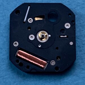 May include: Close-up of a watch movement. The black, octagonal base features various small components, including a gold-colored gear, a copper coil, and several silver-colored screws and pins. The intricate design showcases the internal workings of a timepiece.