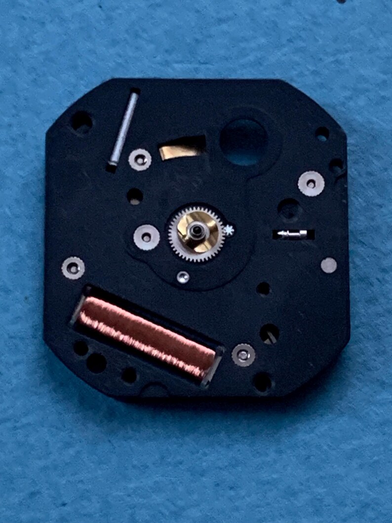 May include: Close-up of a watch movement. The black, octagonal base features various small components, including a gold-colored gear, a copper coil, and several silver-colored screws and pins. The intricate design showcases the internal workings of a timepiece.