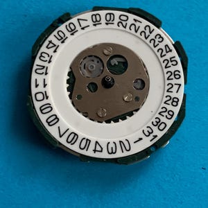 May include: Close-up of a watch movement with a white date wheel and a metal centre. The date wheel has black numbers from 1 to 31. The movement is surrounded by a green plastic ring.