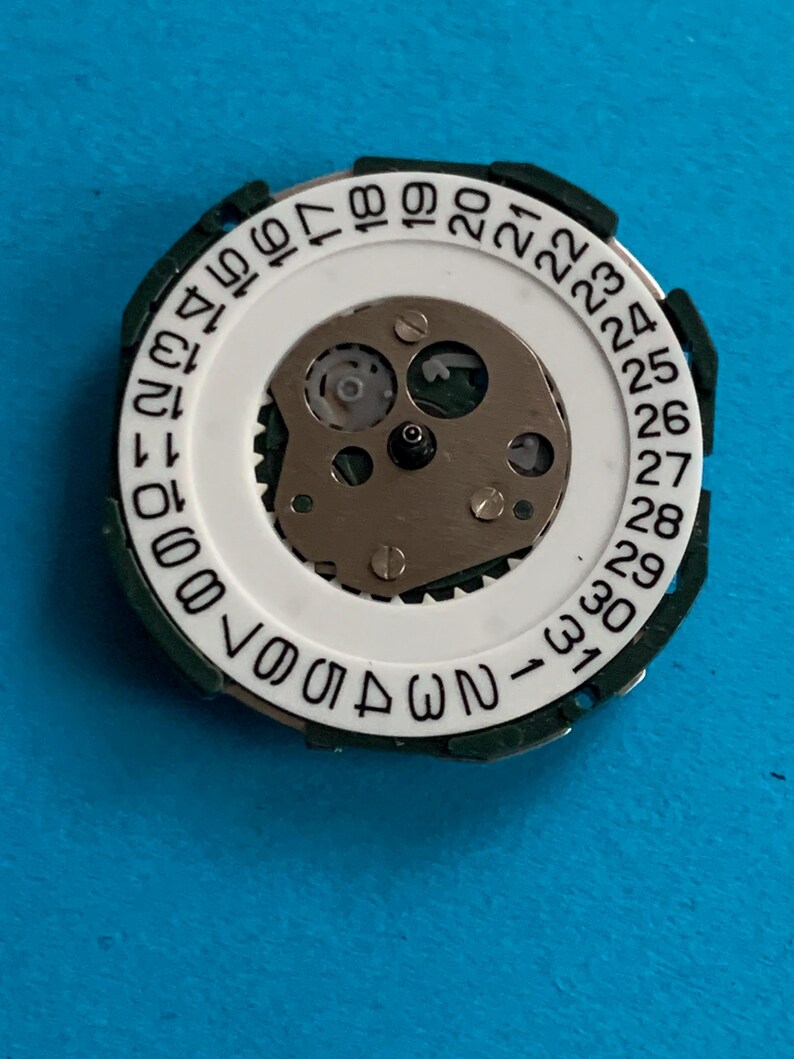 May include: Close-up of a watch movement with a white date wheel and a metal centre. The date wheel has black numbers from 1 to 31. The movement is surrounded by a green plastic ring.