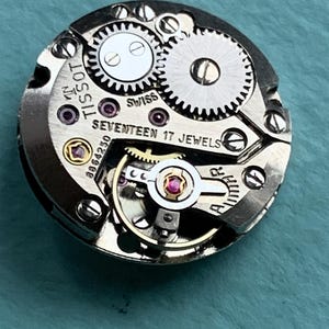 May include: Close-up of a silver watch movement with the text "TISSOT" and "SEVENTEEN 17 JEWELS" visible. The movement has a gold-colored winding stem and a silver-colored rotor.