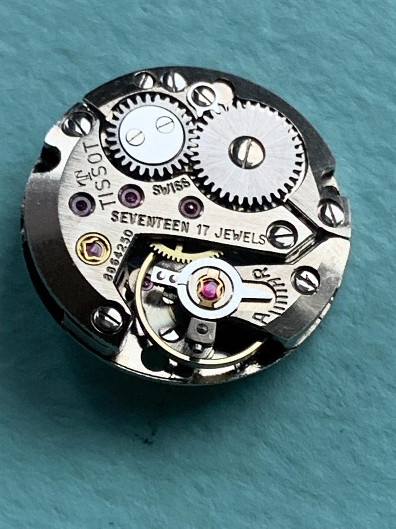 May include: Close-up of a silver watch movement with the text "TISSOT" and "SEVENTEEN 17 JEWELS" visible. The movement has a gold-colored winding stem and a silver-colored rotor.