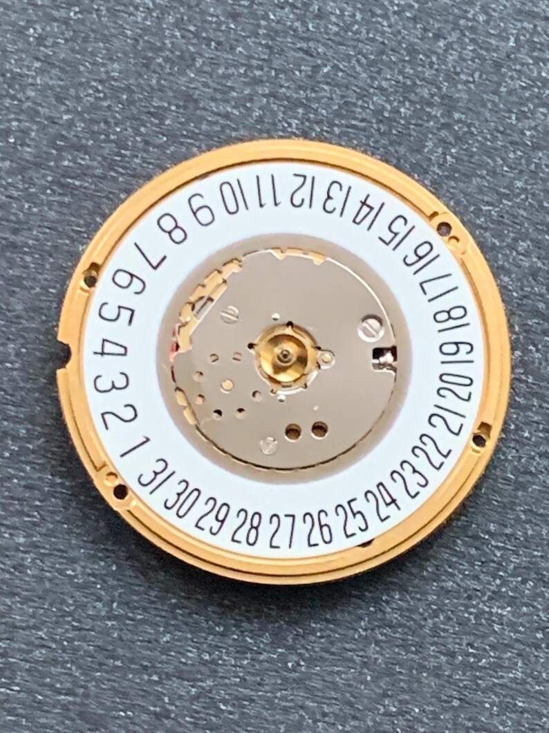 May include: A watch movement with a gold-colored outer ring and a white inner ring. Black numbers from 1 to 31 are printed around the inner ring. The center of the movement has a silver-colored mechanism.