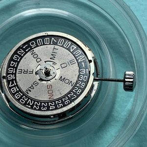 May include: Close-up of a watch movement with a silver and black dial. The dial displays the days of the week in German and numbers for the date. A small screw is attached to the side. The watch movement is inside a clear plastic container.