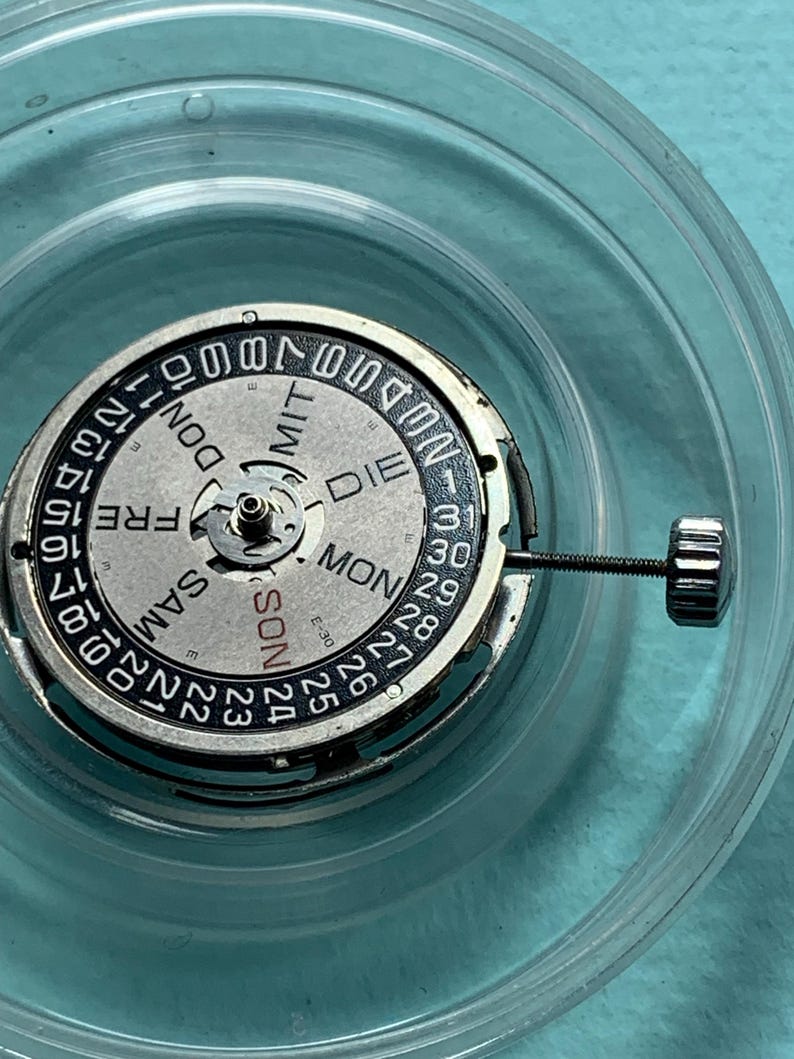 May include: Close-up of a watch movement with a silver and black dial. The dial displays the days of the week in German and numbers for the date. A small screw is attached to the side. The watch movement is inside a clear plastic container.