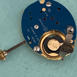 May include: Close-up of a watch movement with a blue circuit board and gold-colored components. The watch mechanism has the text "SWISS SIX 6 JEWELS" and "978.002" printed on it. A small screw is visible.