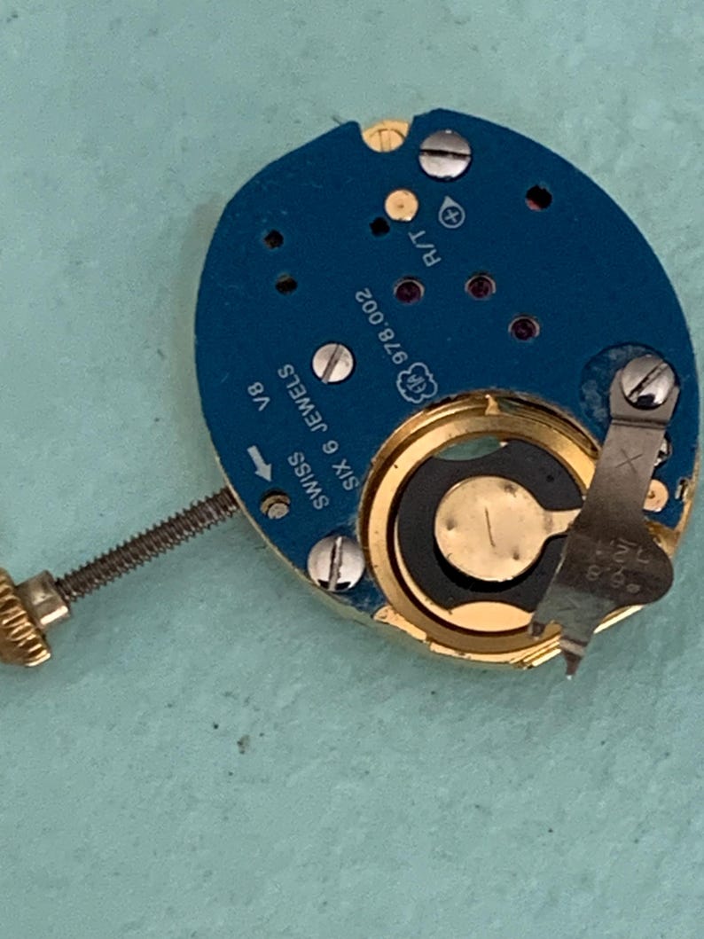 May include: Close-up of a watch movement with a blue circuit board and gold-colored components. The watch mechanism has the text "SWISS SIX 6 JEWELS" and "978.002" printed on it. A small screw is visible.