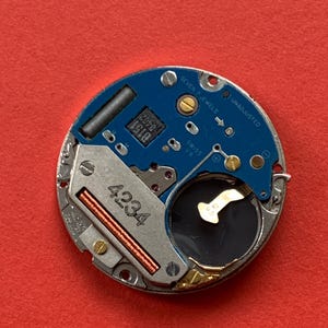 May include: Close-up of a watch movement with a blue circuit board and silver metal components. The text "SEVEN 7 JEWELS UNADJUSTED SWISS V8" is visible. The number "4234" is printed on a silver component.