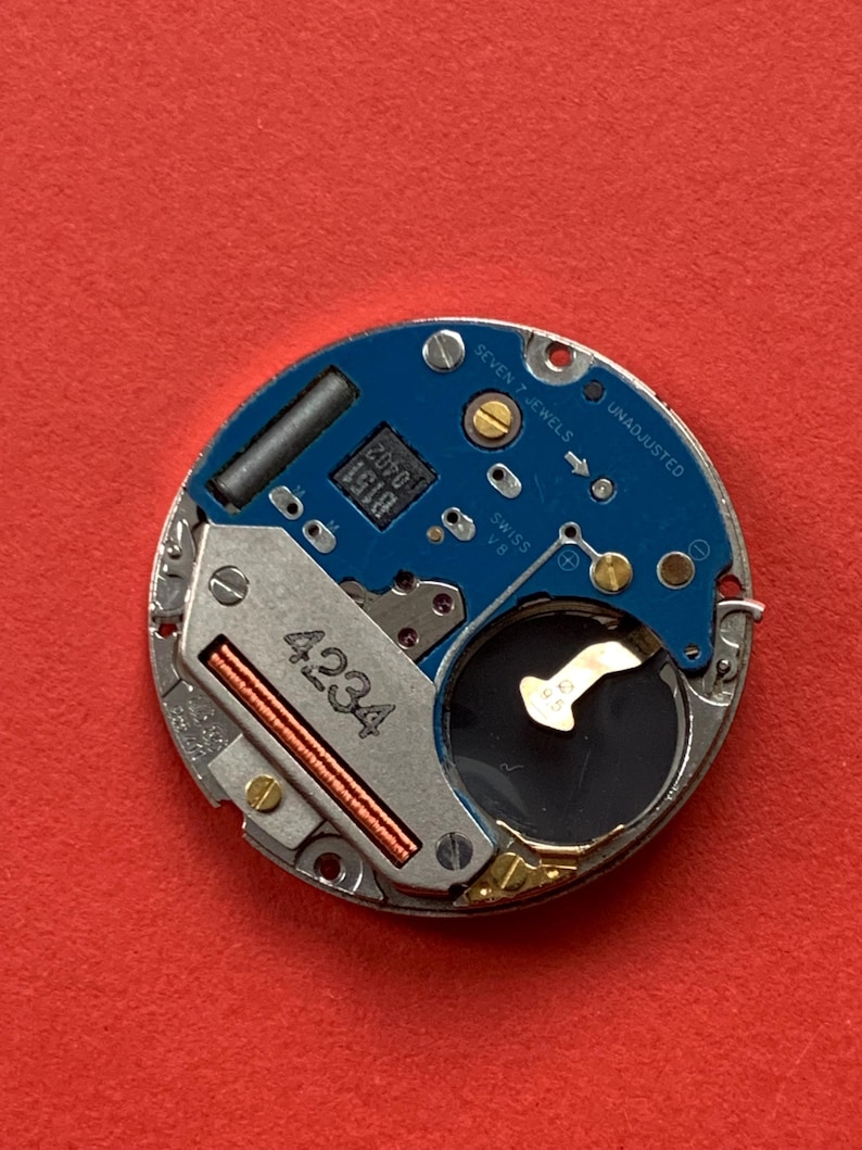 May include: Close-up of a watch movement with a blue circuit board and silver metal components. The text "SEVEN 7 JEWELS UNADJUSTED SWISS V8" is visible. The number "4234" is printed on a silver component.
