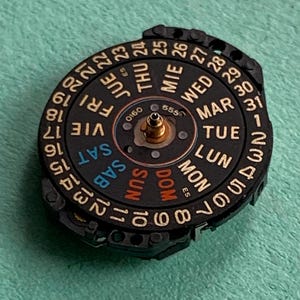 May include: A black watch calendar dial with gold accents, displaying days of the week and numbered dates. The dial features the text "SAT", "SUN", "MON", "TUE", "WED", "THU", and "FRI".