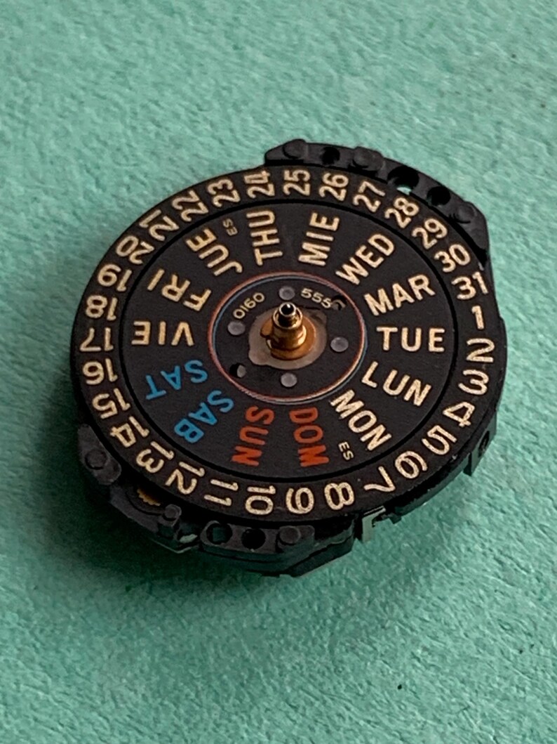 May include: A black watch calendar dial with gold accents, displaying days of the week and numbered dates. The dial features the text "SAT", "SUN", "MON", "TUE", "WED", "THU", and "FRI".