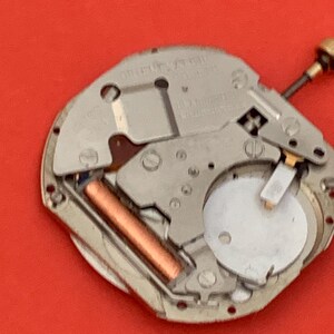 K&ouml;nnte beinhalten: Nahaufnahme eines Uhrwerks mit silberfarbenen Metallkomponenten. Eine kupferfarbene zylindrische Komponente ist sichtbar, zusammen mit einem wei&szlig;en, kreisf&ouml;rmigen Teil. Der Hintergrund ist rot.
