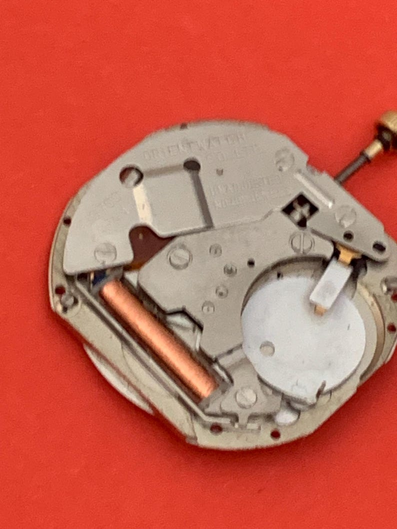 K&ouml;nnte beinhalten: Nahaufnahme eines Uhrwerks mit silberfarbenen Metallkomponenten. Eine kupferfarbene zylindrische Komponente ist sichtbar, zusammen mit einem wei&szlig;en, kreisf&ouml;rmigen Teil. Der Hintergrund ist rot.