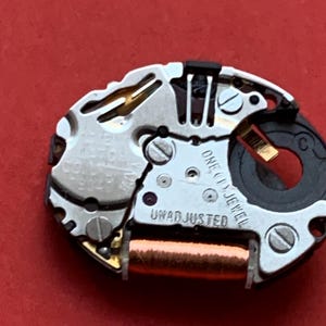 May include: Close-up of a watch movement with silver-colored metal components, screws, and a copper-colored coil. The text "ONE (1) JEWEL" and "UNADJUSTED" are visible. The background is a solid red.