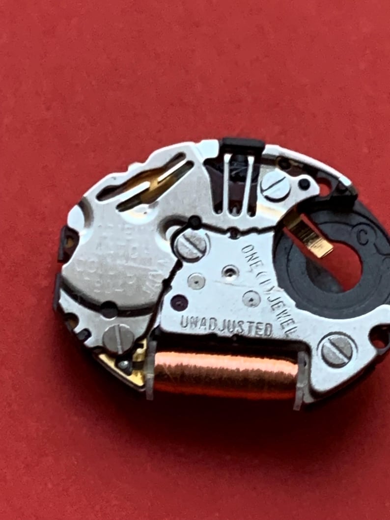 May include: Close-up of a watch movement with silver-colored metal components, screws, and a copper-colored coil. The text "ONE (1) JEWEL" and "UNADJUSTED" are visible. The background is a solid red.