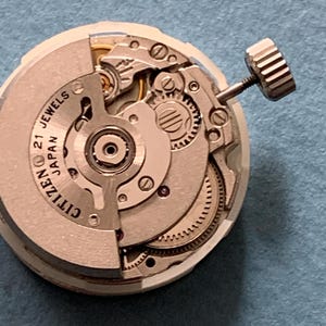 May include: Close-up of a Citizen watch movement with visible gears, screws, and the text "21 Jewels Japan." The watch mechanism is silver-toned with a crown on the side. The intricate details of the watch's internal components are clearly visible.