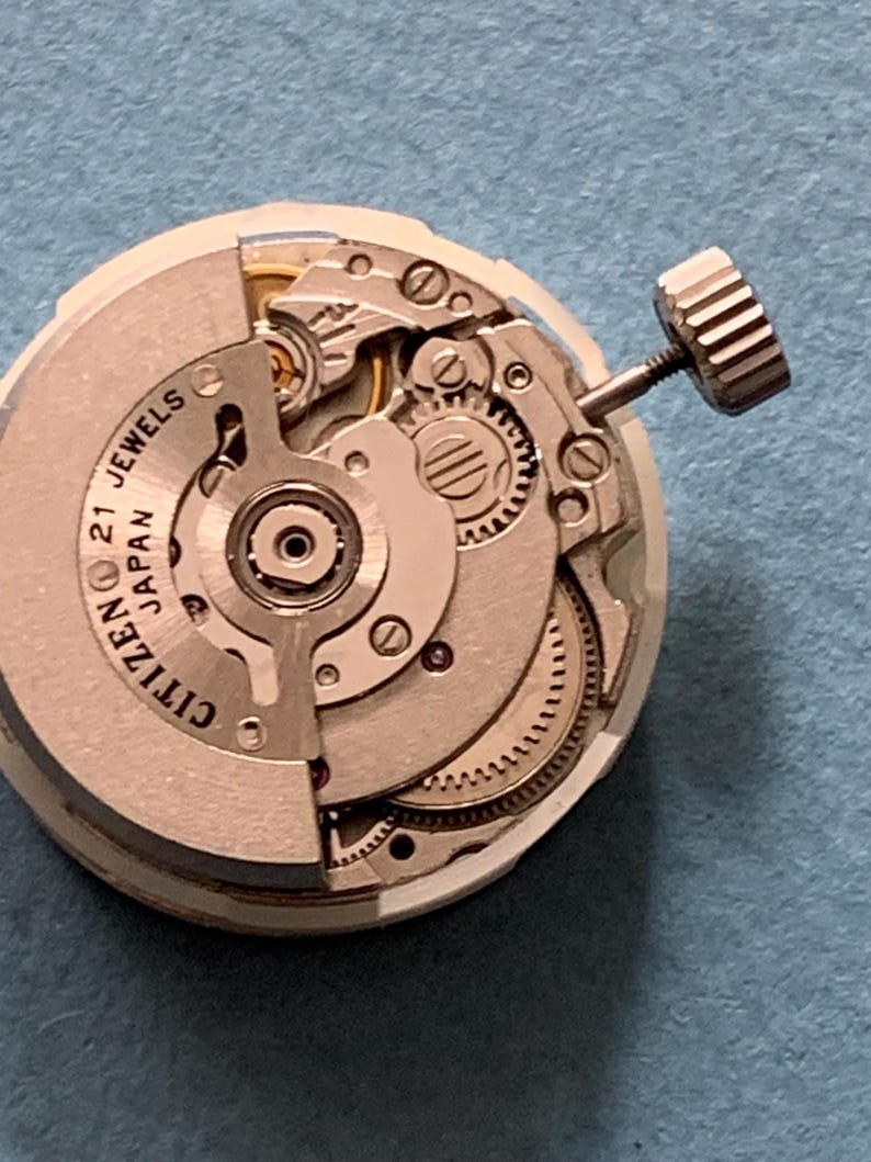 May include: Close-up of a Citizen watch movement with visible gears, screws, and the text "21 Jewels Japan." The watch mechanism is silver-toned with a crown on the side. The intricate details of the watch's internal components are clearly visible.