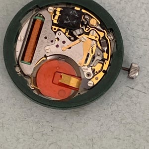 May include: A close-up view of a Citizen watch movement. The watch mechanism is housed in a dark green circular case. The internal components include a copper-colored coil, a red battery, and gold-colored gears and levers. The watch's crown is visible on the right side.